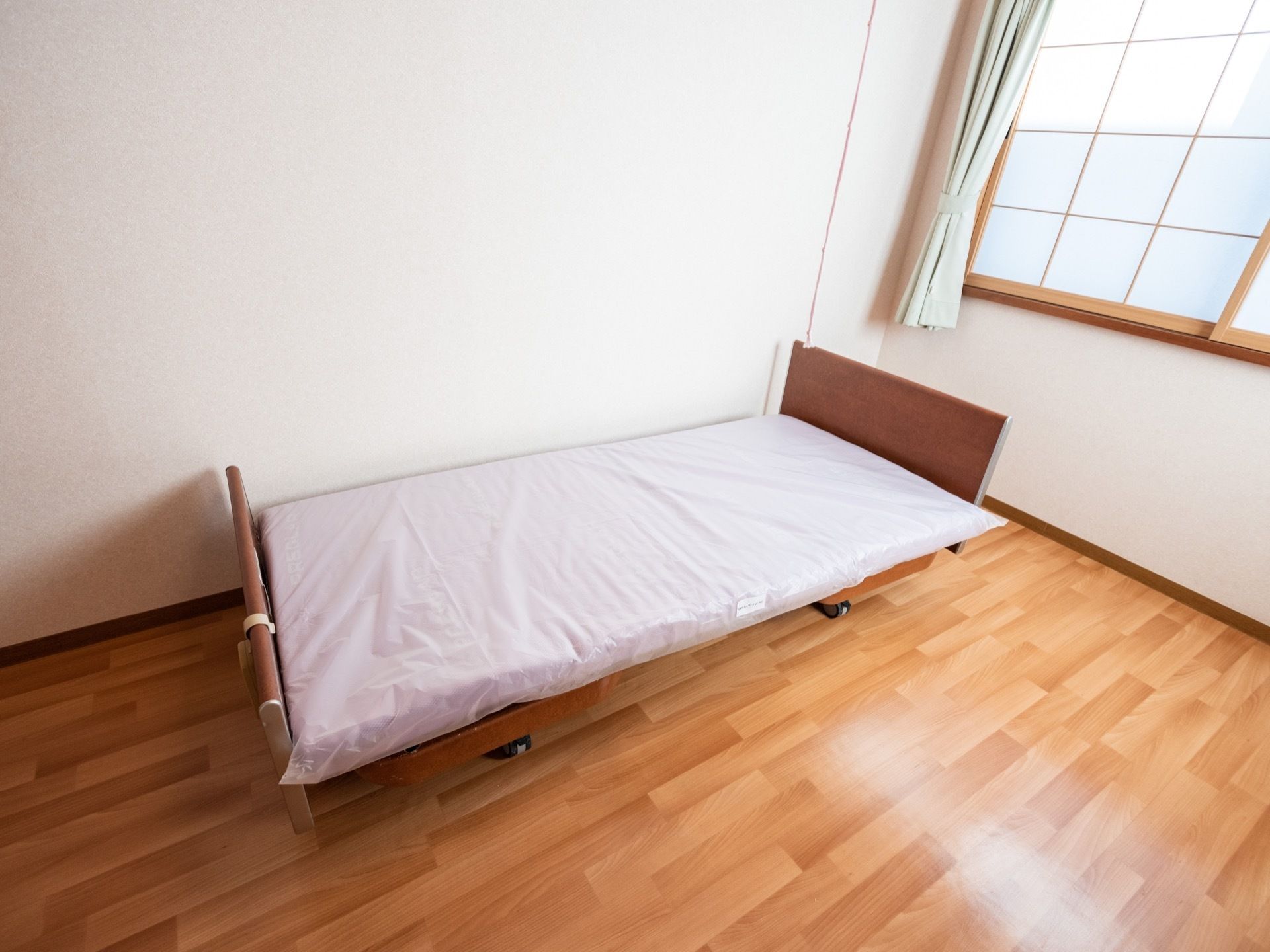 フローリングの床に、白い壁の居室である。壁際に木製のベッドが置かれ、白いマットレスが敷かれている。右奥に障子の引き戸がある。