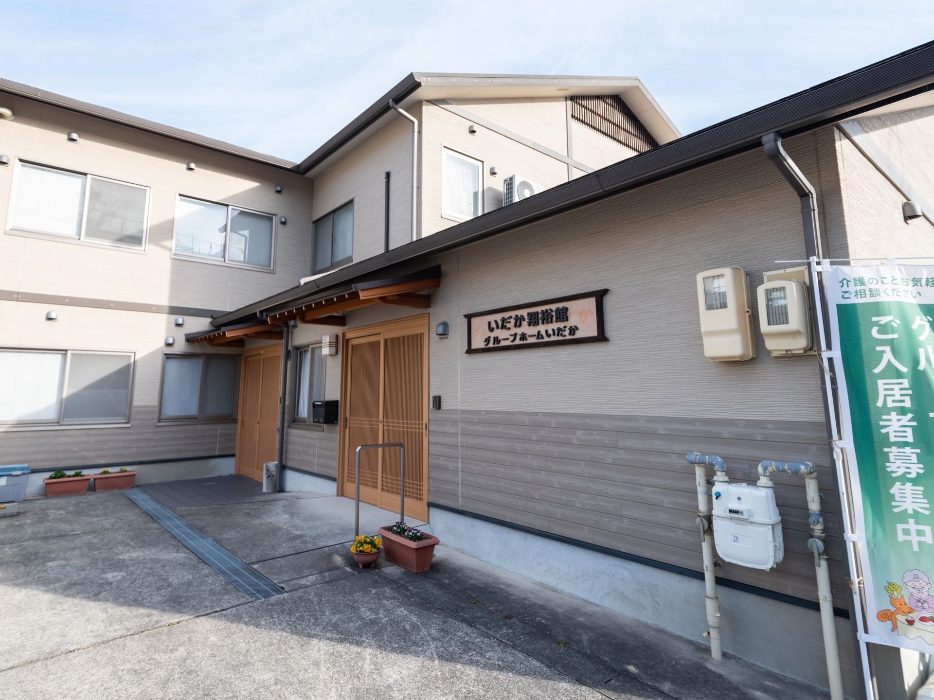 L字型に折れた施設が建っており、２階部分が見えている。中央奥に木製の引き戸の玄関があり、その前の地面に植木鉢が並べられている。