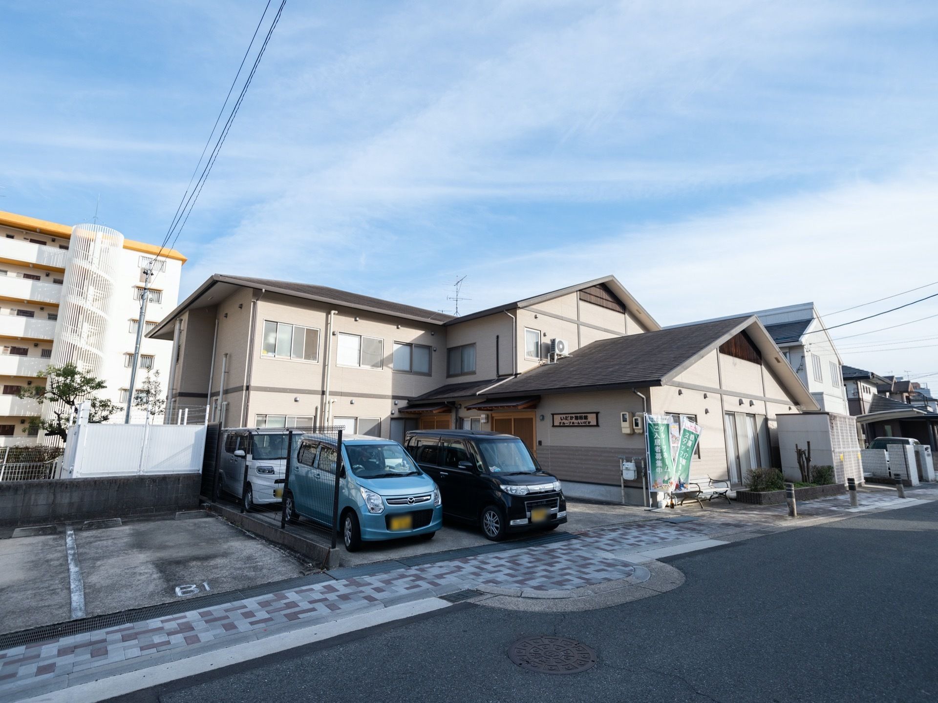 施設前の通りから見た画像である。薄い茶色の外観の施設が中央にある。施設の前は駐車場があり、車が停められている。