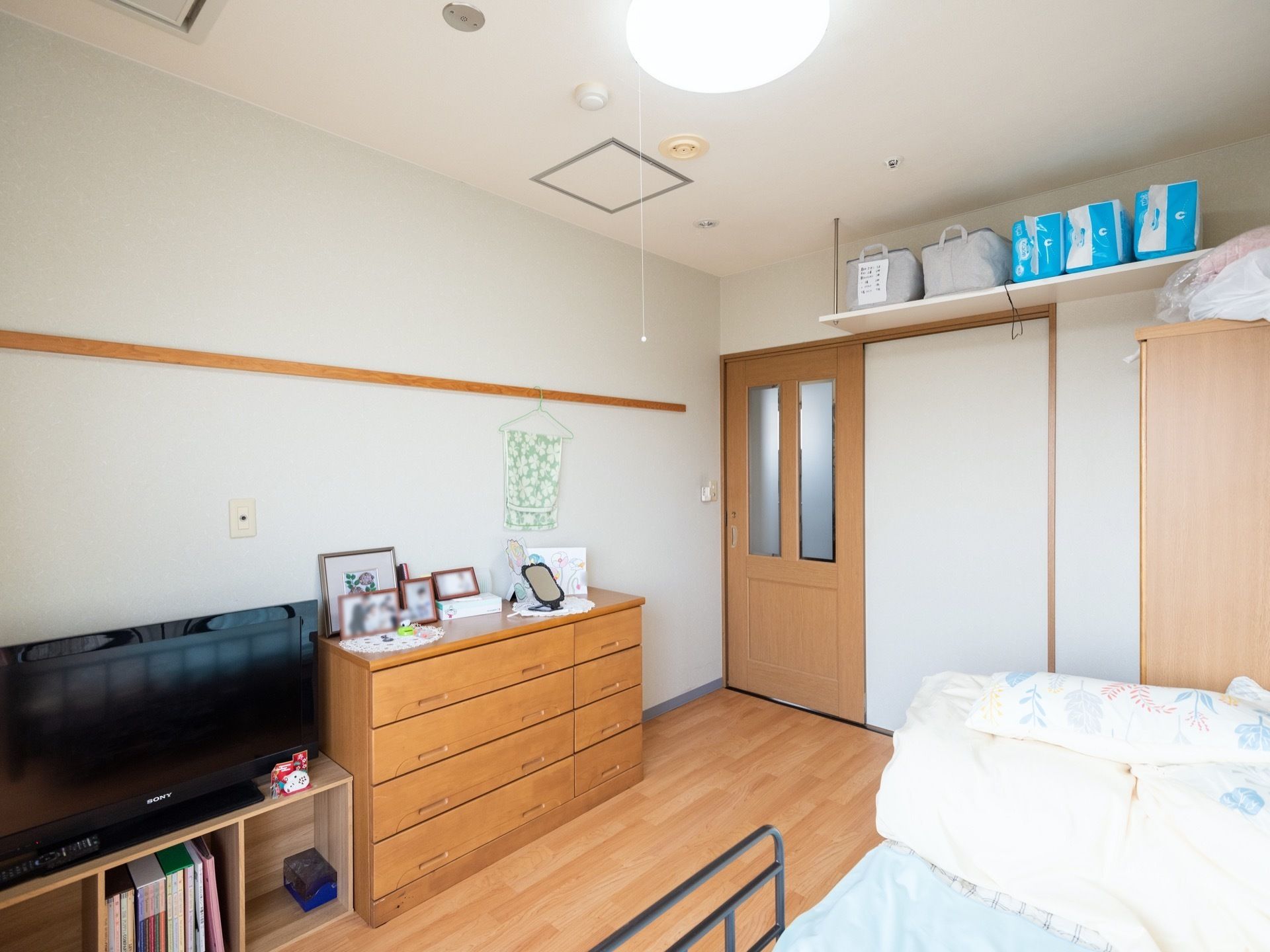 居室の白い壁際には、引出し仕様のカウンター付き整理棚が設置されている。横にはテレビが棚の上に配置されている。