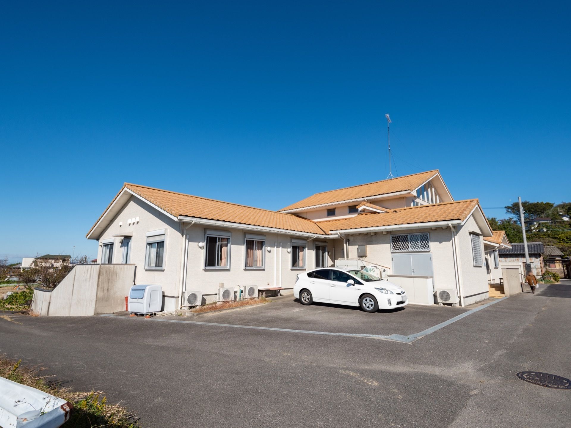 真っ青に晴れ渡った日の平屋建て施設の外観である。完備している駐車場には、1台の白い乗用車が停車している。