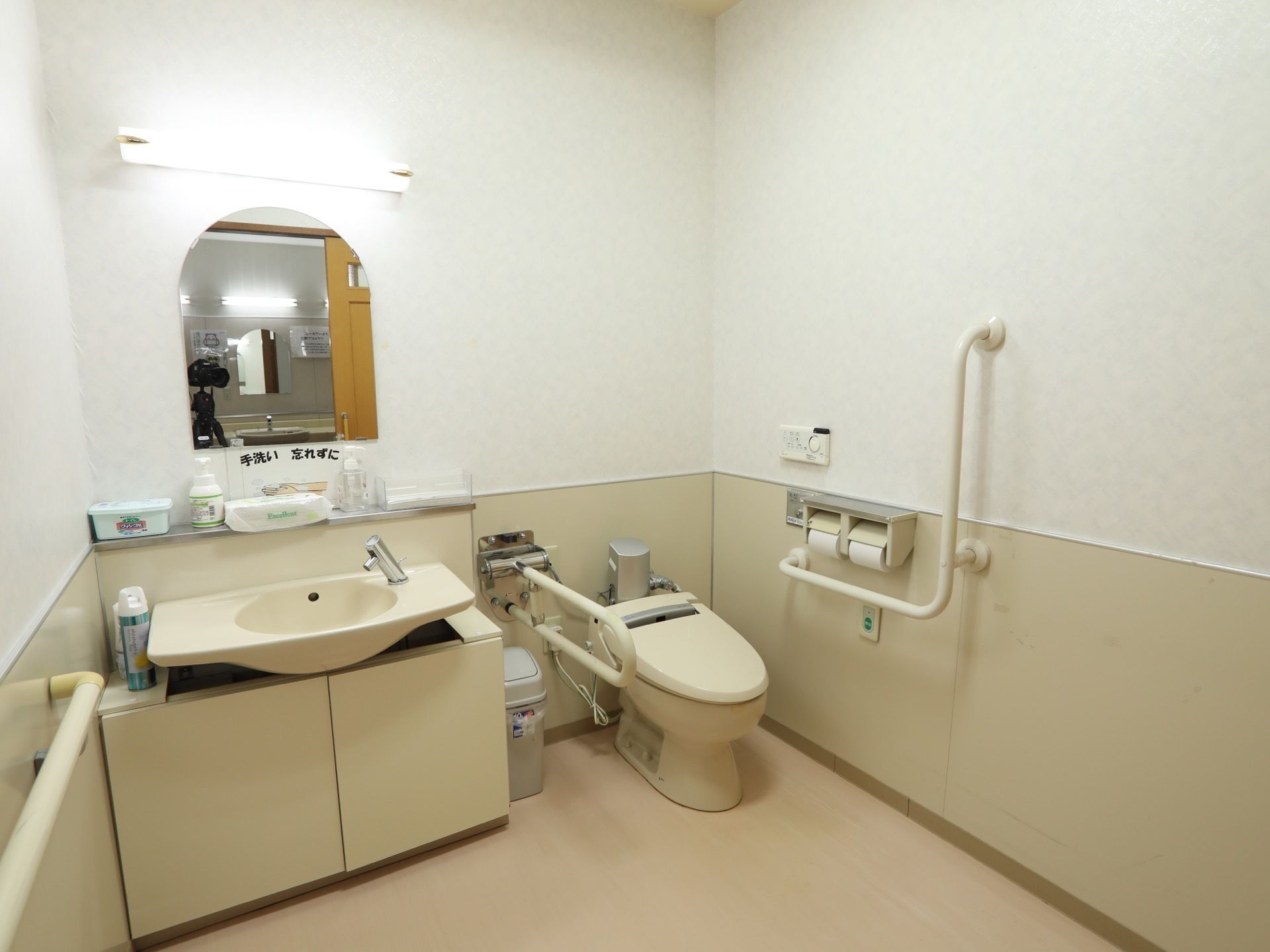 広いトイレである。洗面台と便器が同じ部屋の中にあり、手洗いを促す張り紙が貼られている。便器のそばに緊急時呼び出しボタンが備わっている。