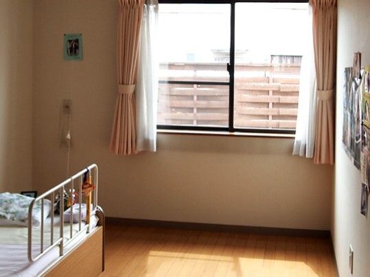 寝具付きのベッドが置かれたフローリングの部屋にはカーテンが付いた窓があり、壁には写真が飾られている。