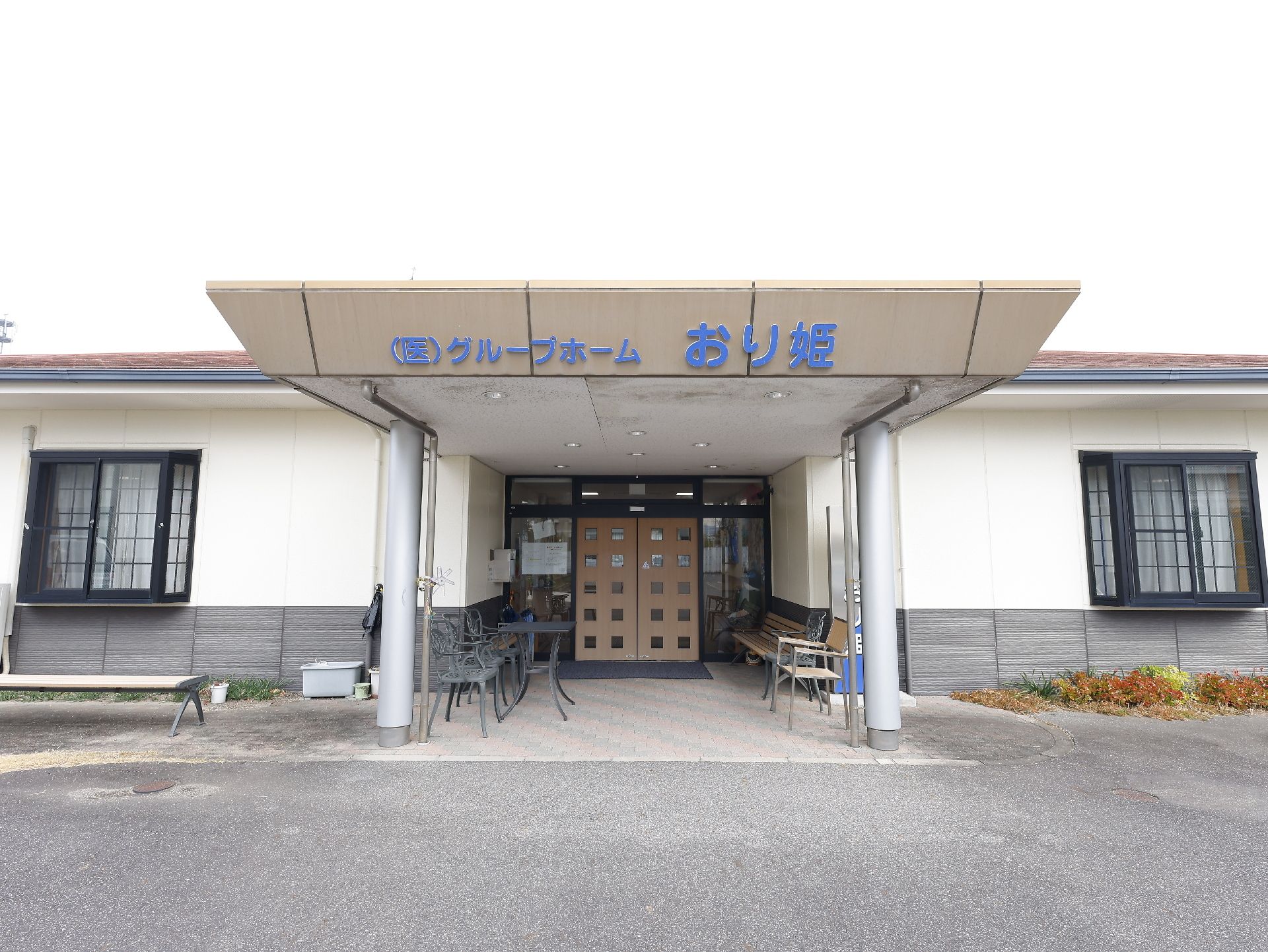 白いゆったりとした服を着た高齢女性が眼鏡をかけた車いすの高齢男性のそばに笑顔で立っている。