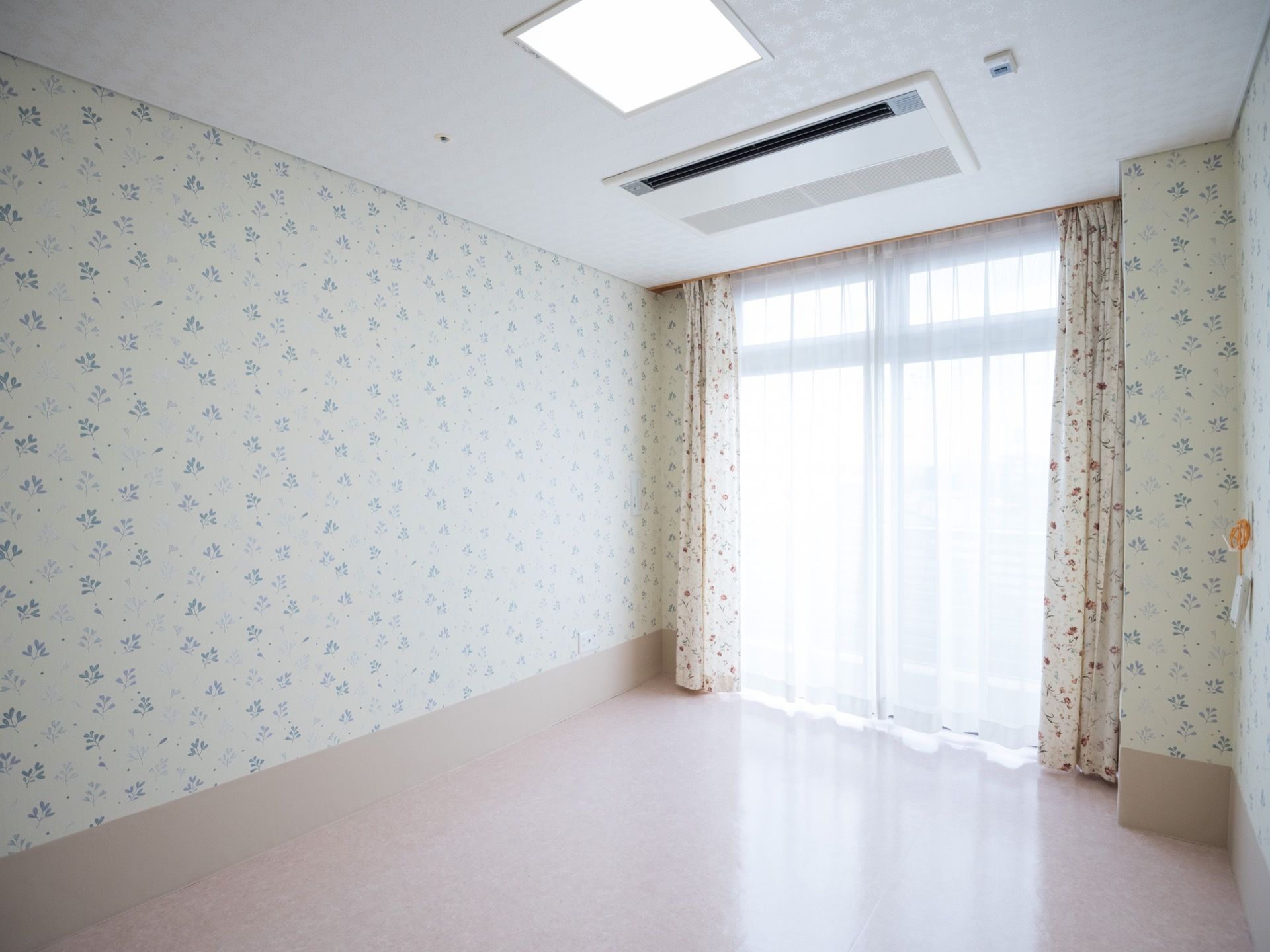 柄の入った壁紙の居室である。大きな窓にはカーテンがかけられている。また、天井には埋め込み式のエアコンが設置されている。