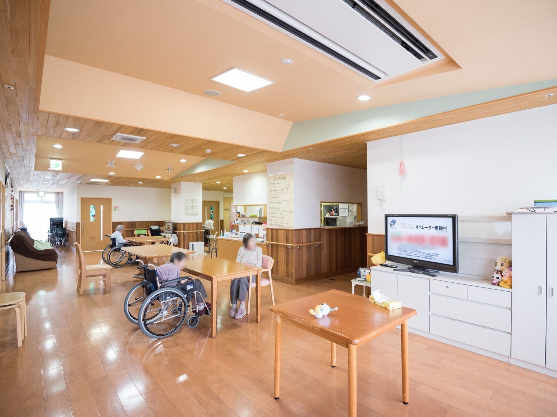 白色の棚の上にテレビが１台あり、入居者様がテレビを見ている様子である。奥の方には、茶色のカバーがかけられたソファも置かれている。