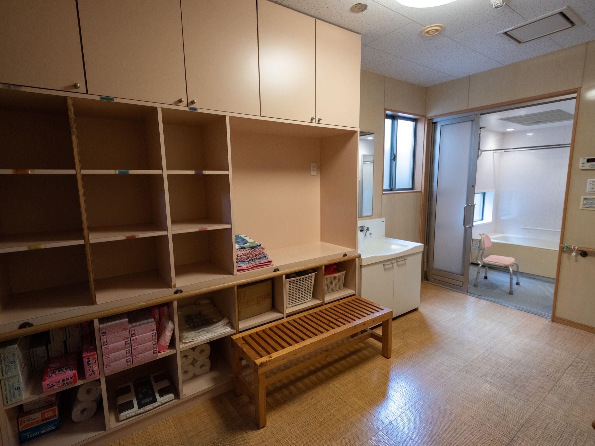 壁側に収納棚があり、タオルやゴミ袋、体重計など、さまざまな物が置かれている。棚の前にベンチが１台設置されている。