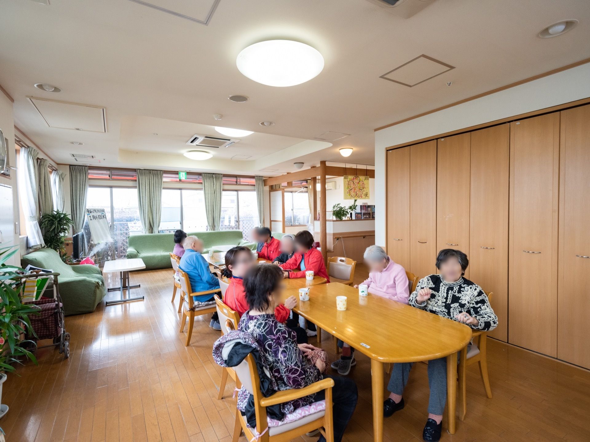 テーブル席にスタッフと入居者様が座っており、楽しそうに談笑している。窓側には、ソファが設置されている。