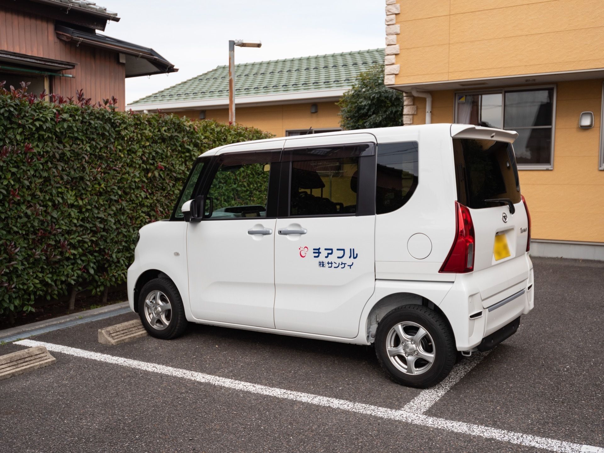 施設前の路面はアスファルトで舗装されており、駐車スペースになっている。施設名が側面に書かれた白い車が停まっている。