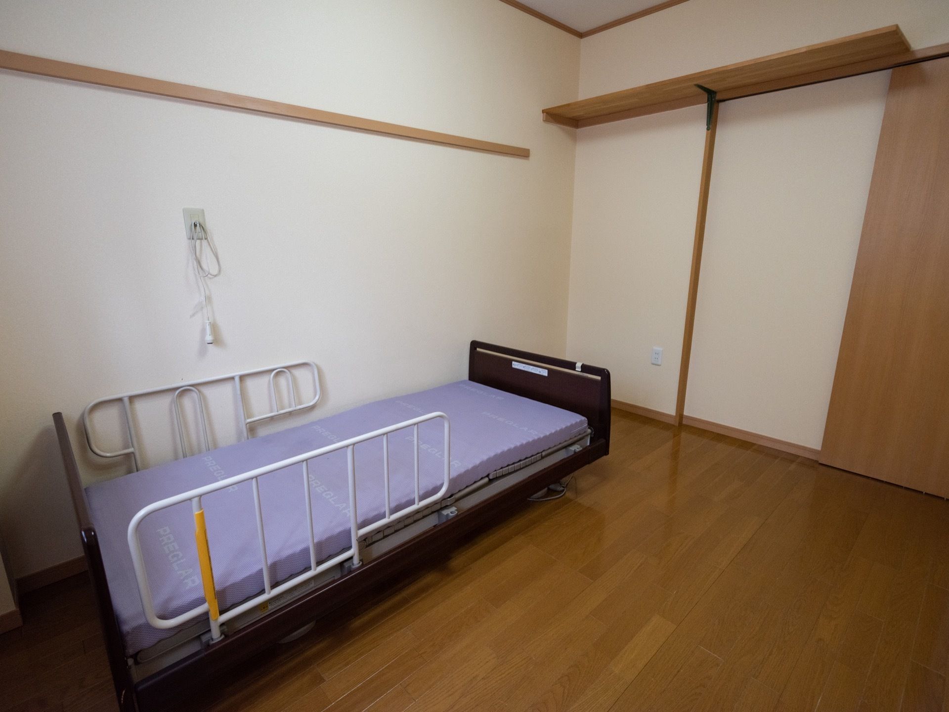 壁側にベッド柵の取り付けられた電動ベッドが１台置かれている。ベッド近くの壁にはナースコールが設置されている。