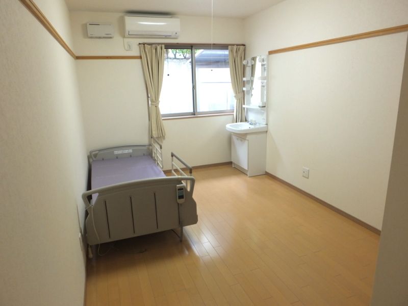 寝具のない介護ベッドやエアコン、洗面所などが設置されたフローリングの部屋。