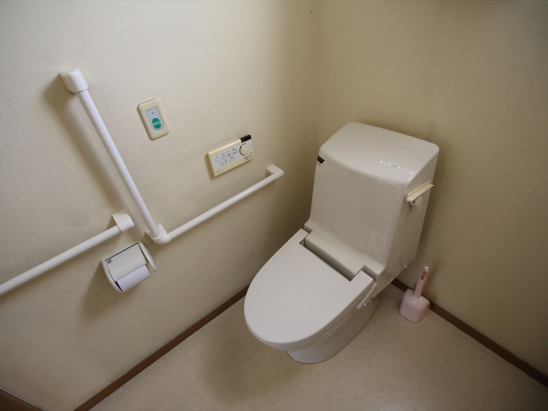白い壁のトイレがある。温水洗浄便座が付いており、壁には手すりがある。非常用のブザーが設けられている。