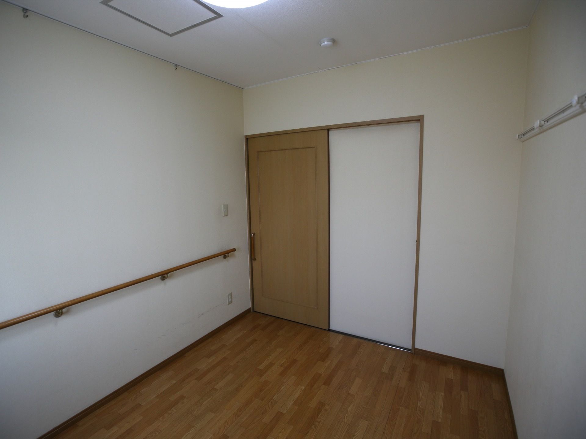 白い壁とダークブラウンの床の居室がある。引き戸のドアが付いており、壁には木製の手すりがある。