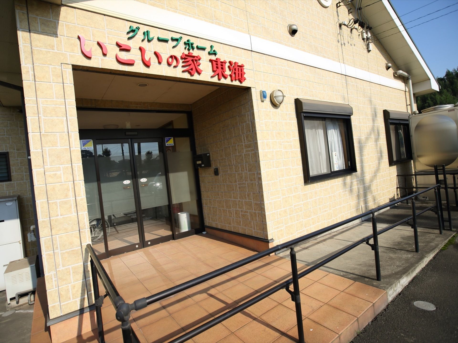 ベージュのタイルの壁に囲まれた玄関がある。両開きのガラス張りの自動ドアが付いている。玄関へはスロープと手すりがある。