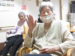 付き添いの高齢男性と車いすの高齢女性が笑顔で屋外で過ごしている。両サイドには若い男女のスタッフがいる。
