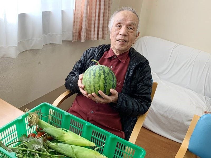 椅子に座った高齢男性がスイカを両手で持っている。テーブルの上にはトウモロコシなどもある。