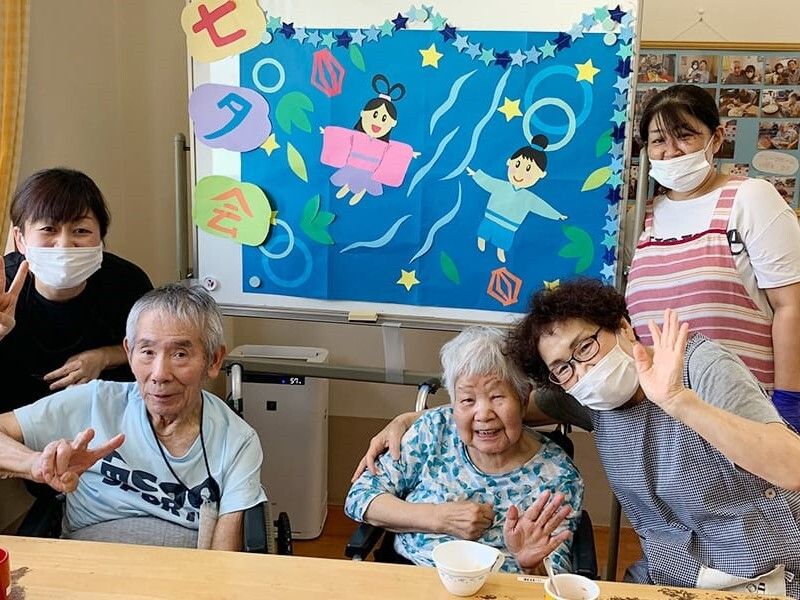 「七夕会」とカラフルな文字で飾られたホワイトボードには彦星と織姫も飾られ、入居者とスタッフが笑っている。