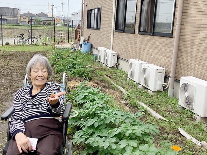 白髪の高齢女性が車いすに座り、菜園の前でピースサインをしている。たくさんの野菜が植えられている。