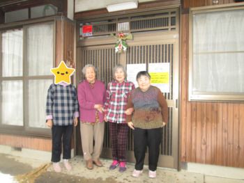 玄関は引き戸で家庭的な雰囲気の民家。しめ縄が飾られ、高齢女性たちが腕を組んで微笑んでいる。