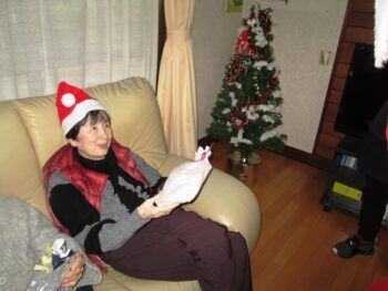 ベージュのソファに座ってサンタ帽をかぶっている高齢女性。プレゼントを受け取り、微笑んでいる。