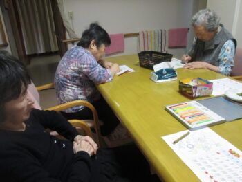 塗り絵に励んでいる3名の高齢女性。手前に座っている女性は鉛筆を持たず、紙を眺めている。