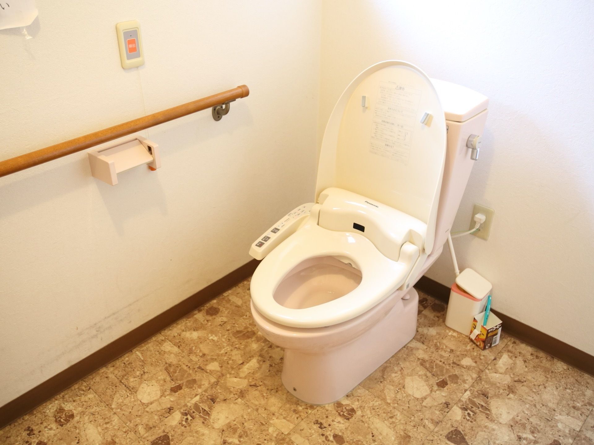 トイレの白い壁には、手すりや緊急呼び出しブザー、ペーパーホルダーが設置されている。温水洗浄便座が完備されている。