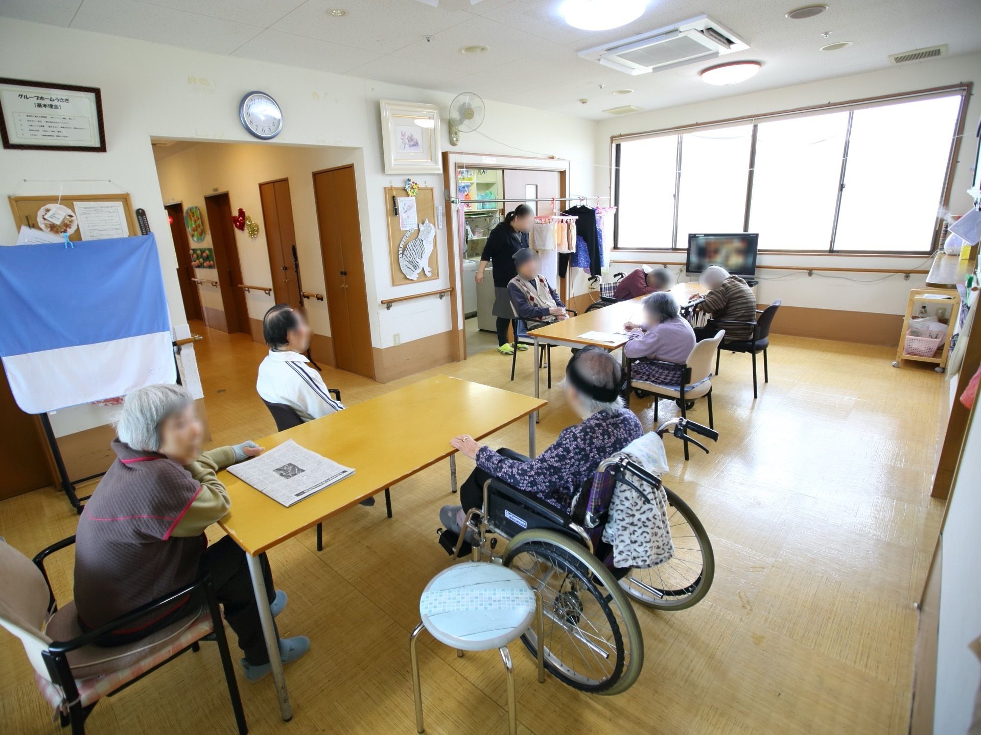 窓が設置された共有スペースには、テーブルが２つ設置されている。車椅子をご利用の方や椅子に腰掛けられている入居者様の姿が見える。