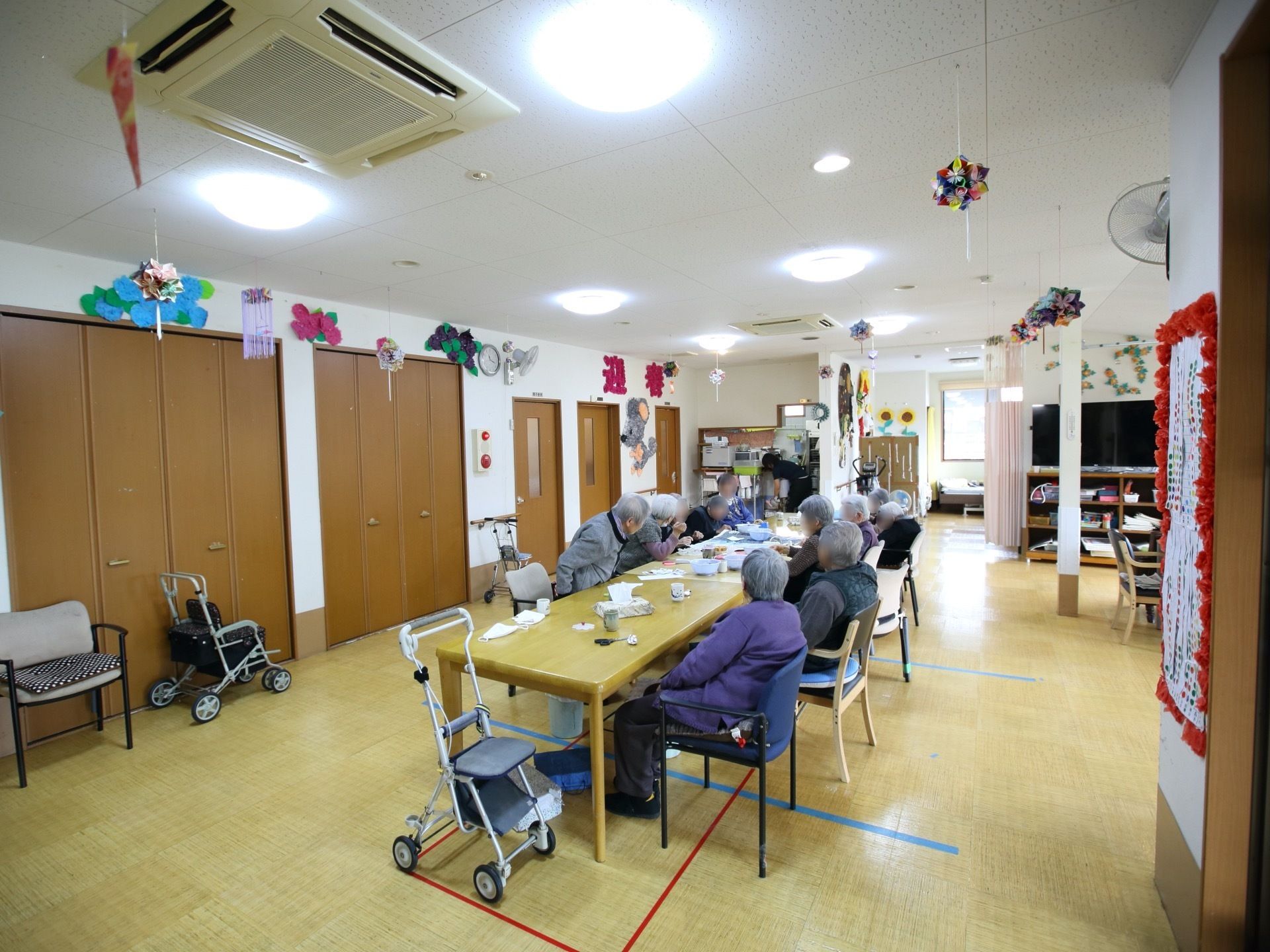 共有スペースには、季節の飾り付けが壁に施されている。中央に１列に配置されたテーブルセットには、入居者様皆さまが席に着かれている。