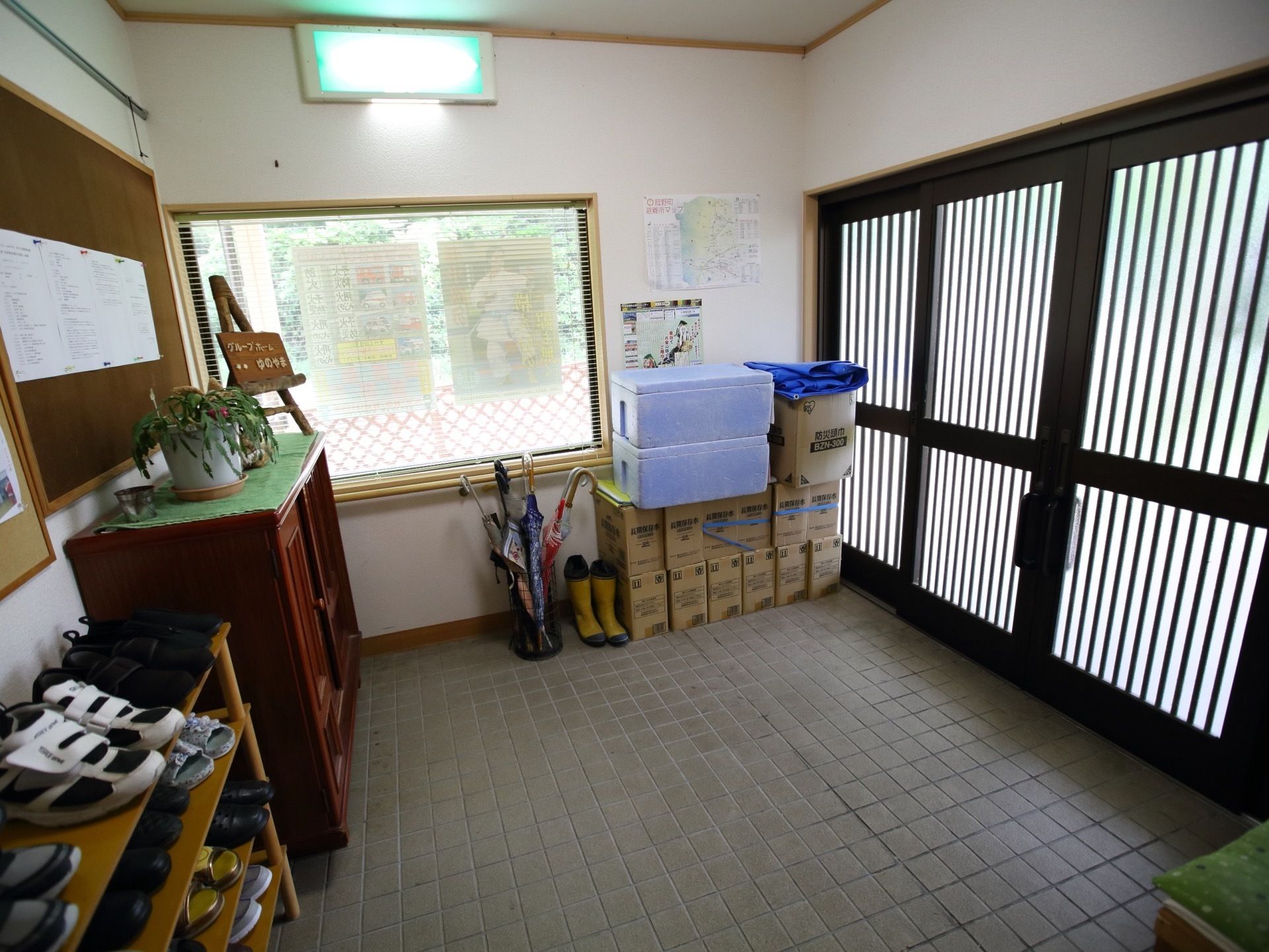 灰色のタイルが敷き詰められ、広々とした玄関である。茶色の棚の上には植物が置かれている。下駄箱や傘も備え付けられている。