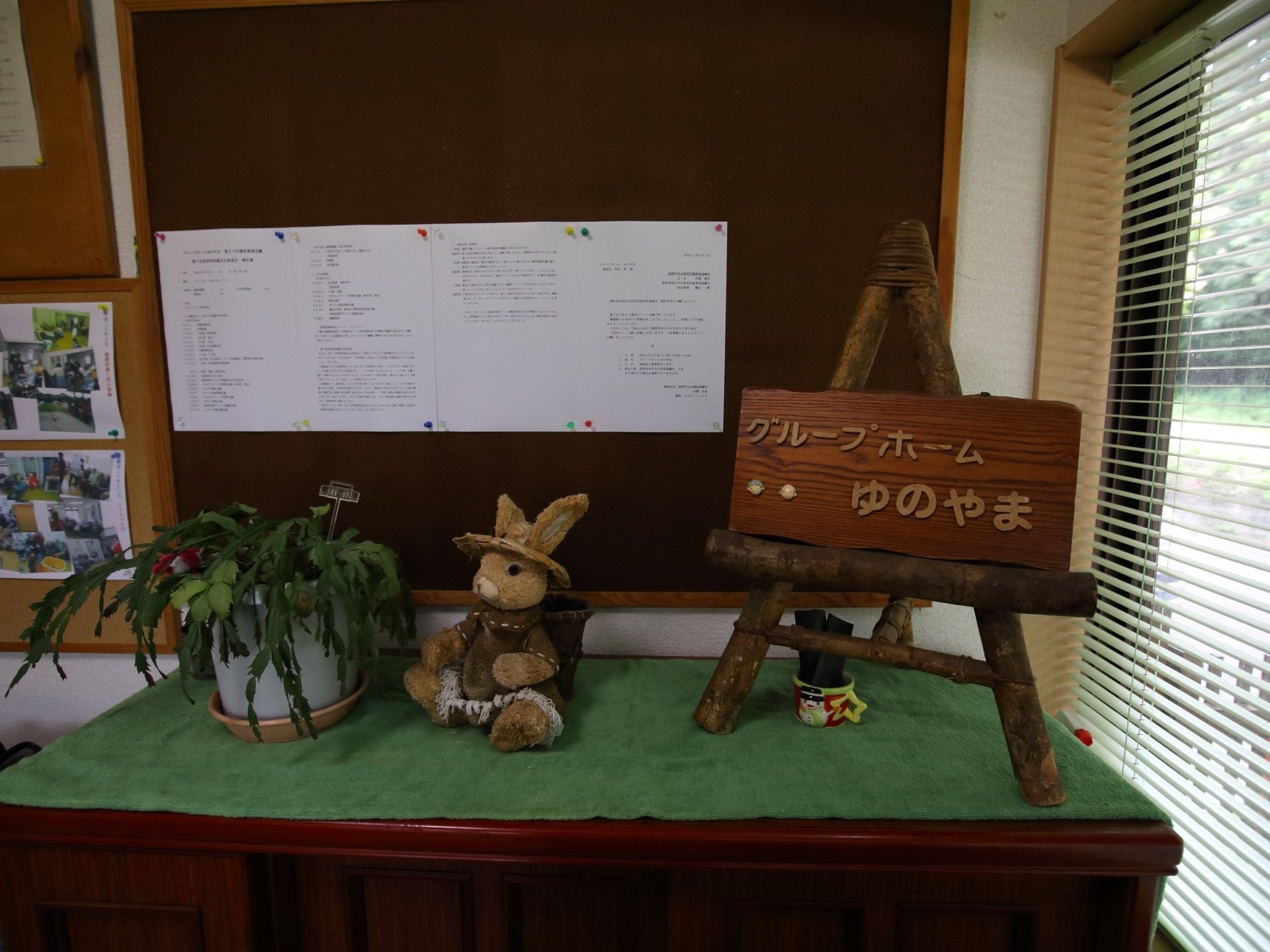 緑の布の上にグループホームの看板と、うさぎの置物と植物が設置されている。後ろのボードには紙や写真が貼られている。