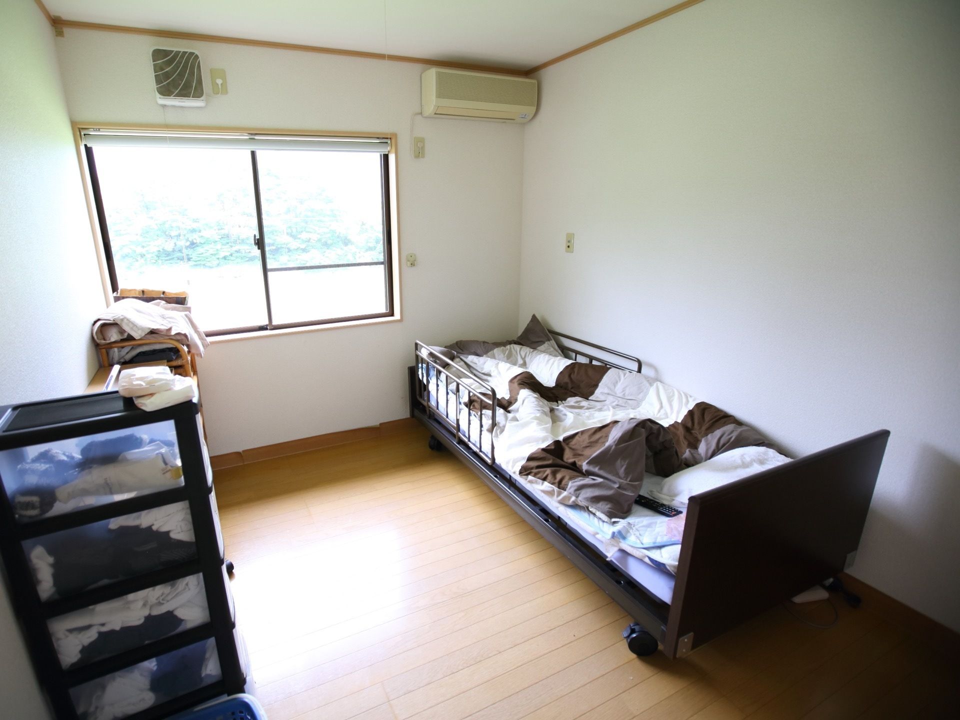 白色の壁と茶色のフローリングで、落ち着いた色合いの内装の居室である。エアコンや棚、ベッドが置かれている。