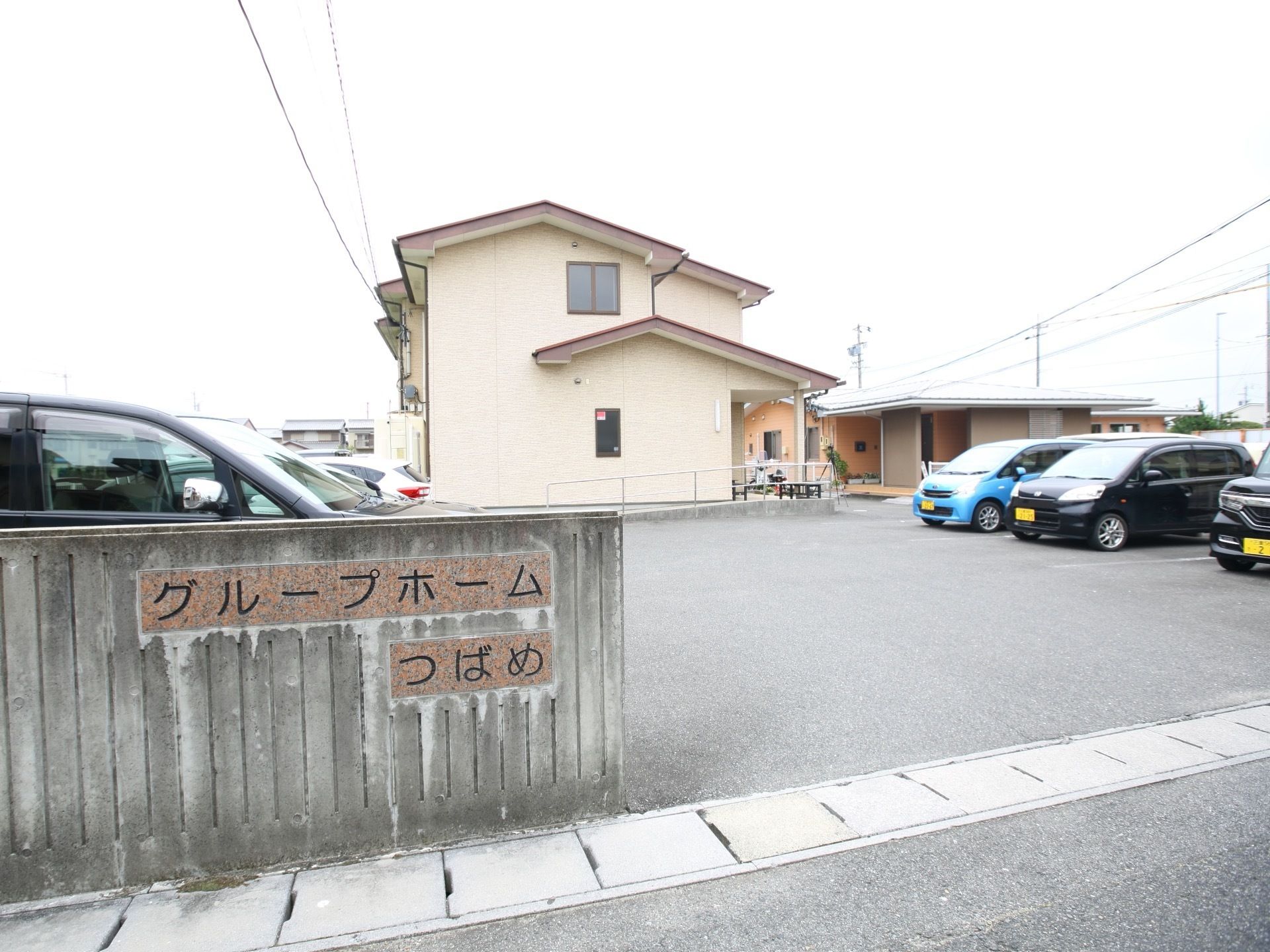 淡いベージュの外壁で、２階建ての建物である。スロープが設置されている。駐車スペースが広く設けられている。