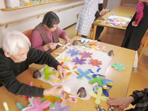 食堂のテーブルで、入居者様が色紙で大きな花を作成している。その花を大きな白い模造紙に貼っている。花は色数が多く、華やかである。
