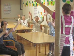 入居者様がダイニングの椅子に座り、両手を高く上げて体操をしている。前にはエプロン姿のスタッフが立って、指導をしている。