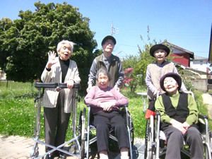 青空の下で、入居者様がポーズを取っている。車椅子に乗っている方や歩行器を使っている方もいる。後ろに大きな木や白い花が見える。