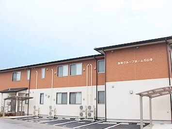 2階建ての建物はベージュとブラウンの外壁で、建物の前には駐車スペースがあり、玄関には屋根が付いている。