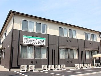 グレーとベージュの2階建ての建物の前には駐車スペースがあり、外壁にはホーム名や併設事業所名が書かれている。