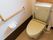 トイレに、薄緑色の洋式便座がある。壁には大きなL字型の手すりが取り付けられていて、その下にトイレットペーパーホルダーがある。
