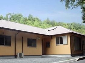 緑に囲まれた場所に建てられているベージュ系の外壁の平屋建ての建物はグレーの屋根が付いている。