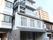 街中にある白を基調とした建物。両サイドにも民家やビルのようなものがあり、1階部分は駐車場になっている。