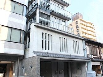 縦に細長い建物は白を基調としていて、1階部分は駐車場。周りには民家やビルもある。
