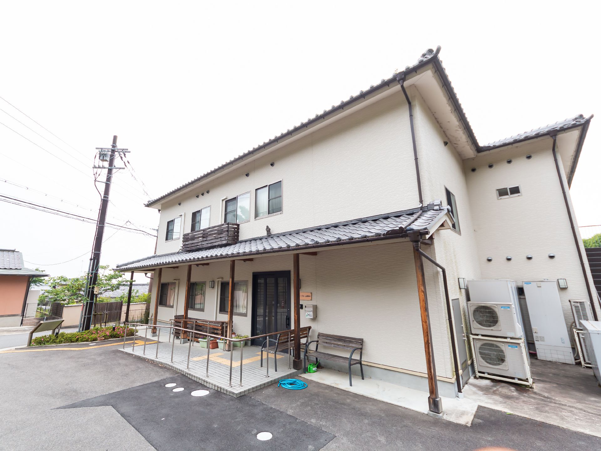 グレーの瓦屋根が付いた白壁の2階建ての建物の周辺には緑が多く、玄関には手すり付きのスロープが付いている。