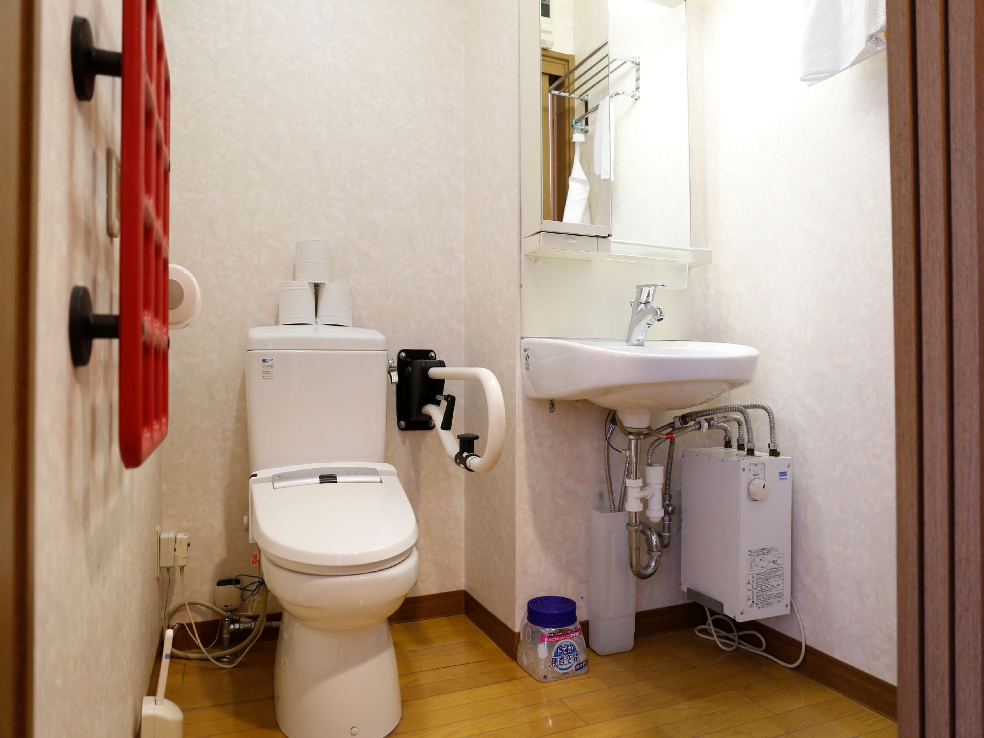 白調の壁に木目調の床。便器の傍横には手すりがある。その隣に洗面台が設置され、上には細長い鏡。便器後方に予備のトイレットペーパーが置いてある。
