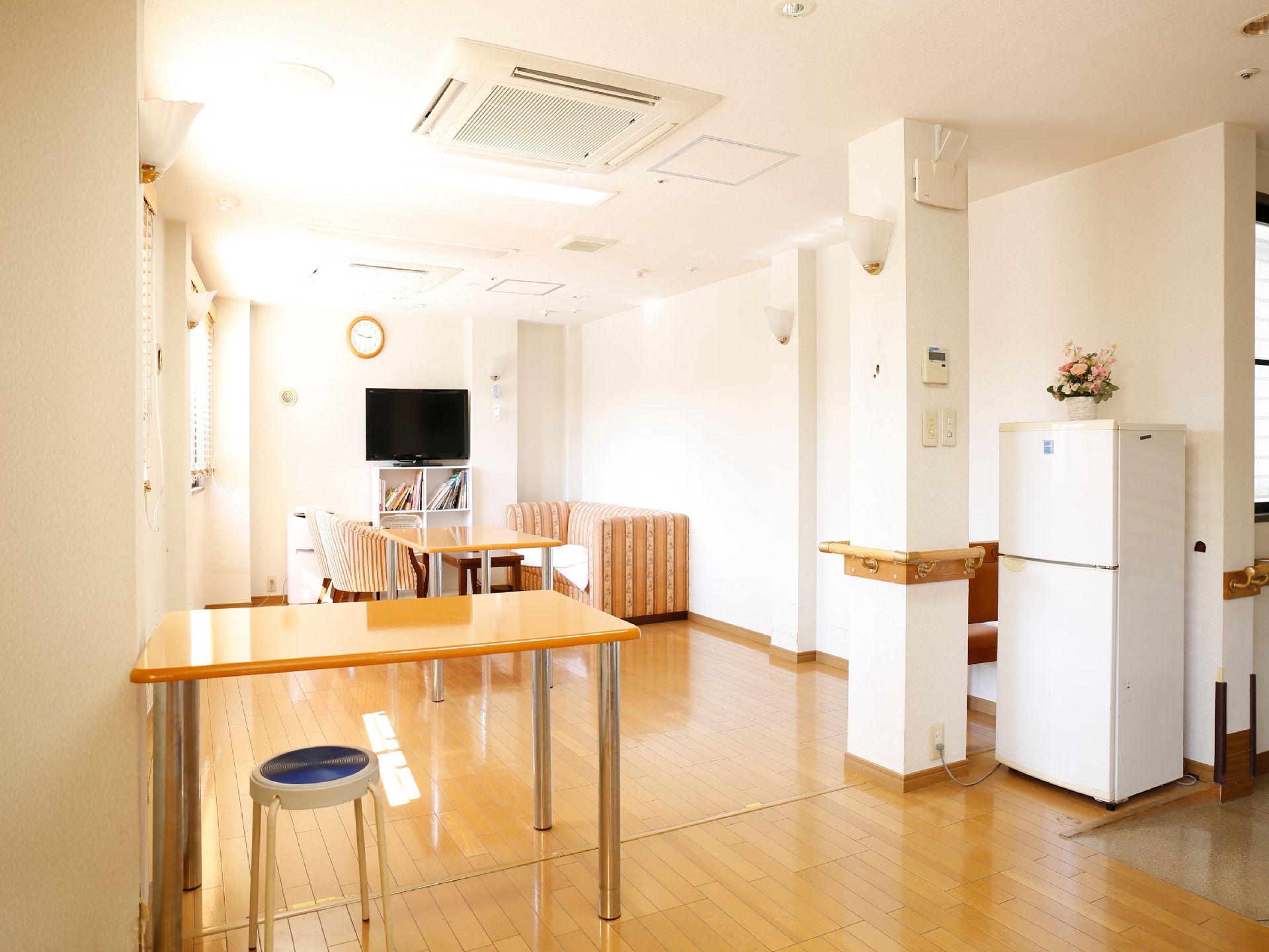 白色の壁と天井、木目調の床。奥には本棚の上にテレビが置いてあり、その前には、テーブルとソファが置かれている。手前には冷蔵庫がある。