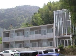 グレーの2階建ての建物の周りには緑が多く、奥には大きな山もある。建物の前には駐車スペースがあり、車が止まっている。