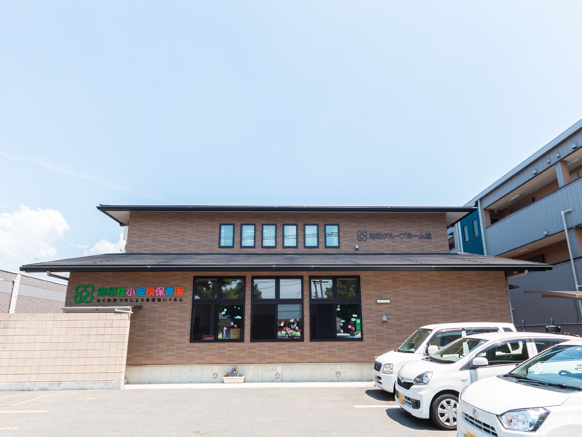 ベージュの2階建ての建物の玄関には屋根が付き、カラフルな文字でホーム名が書かれている。窓もたくさんついている。