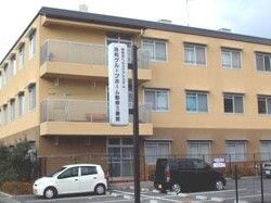 ベージュの3階建ての建物には窓が多く、建物の前には駐車スペースがあり、車が2台止められている。
