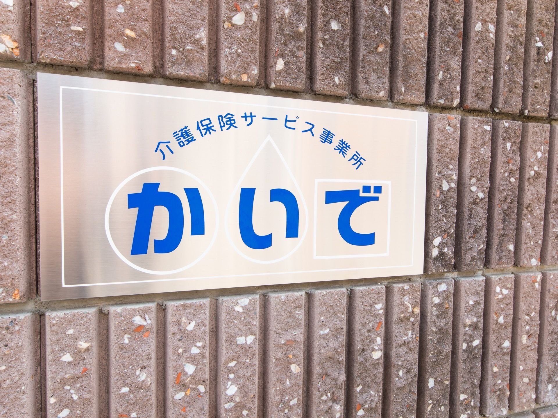 茶色い縦向きのレンガの上に、銀色のプレートが設置してある。白い枠線の中に、青い文字が一文字ずつ入っている。