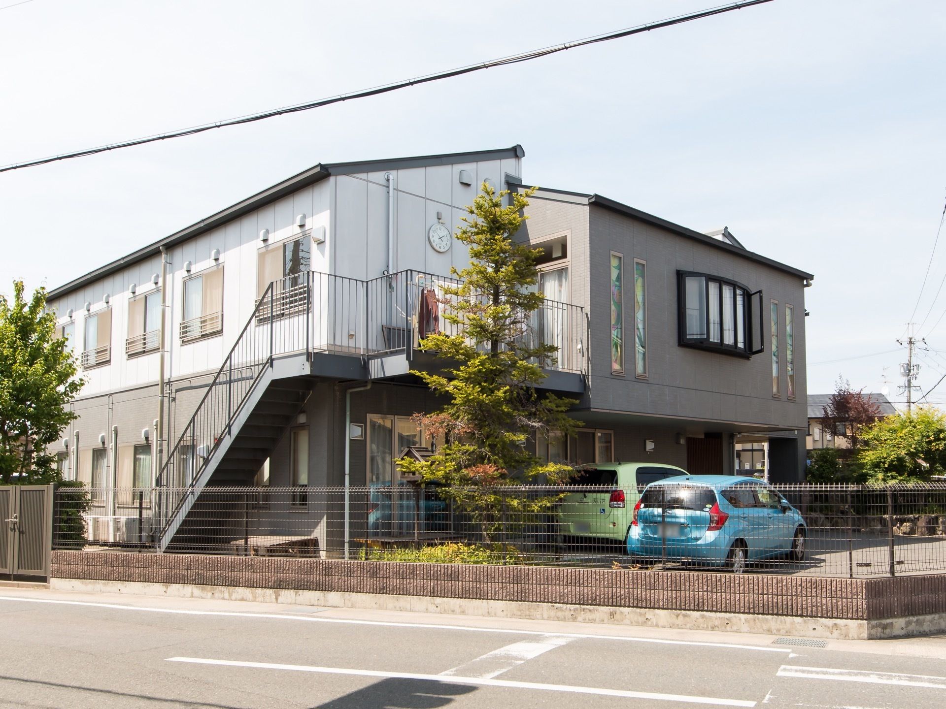 ２階建ての建物は、道路側に外階段が取り付けてある。階段上の壁に時計が設置してあり、駐車場からも時間がわかる。