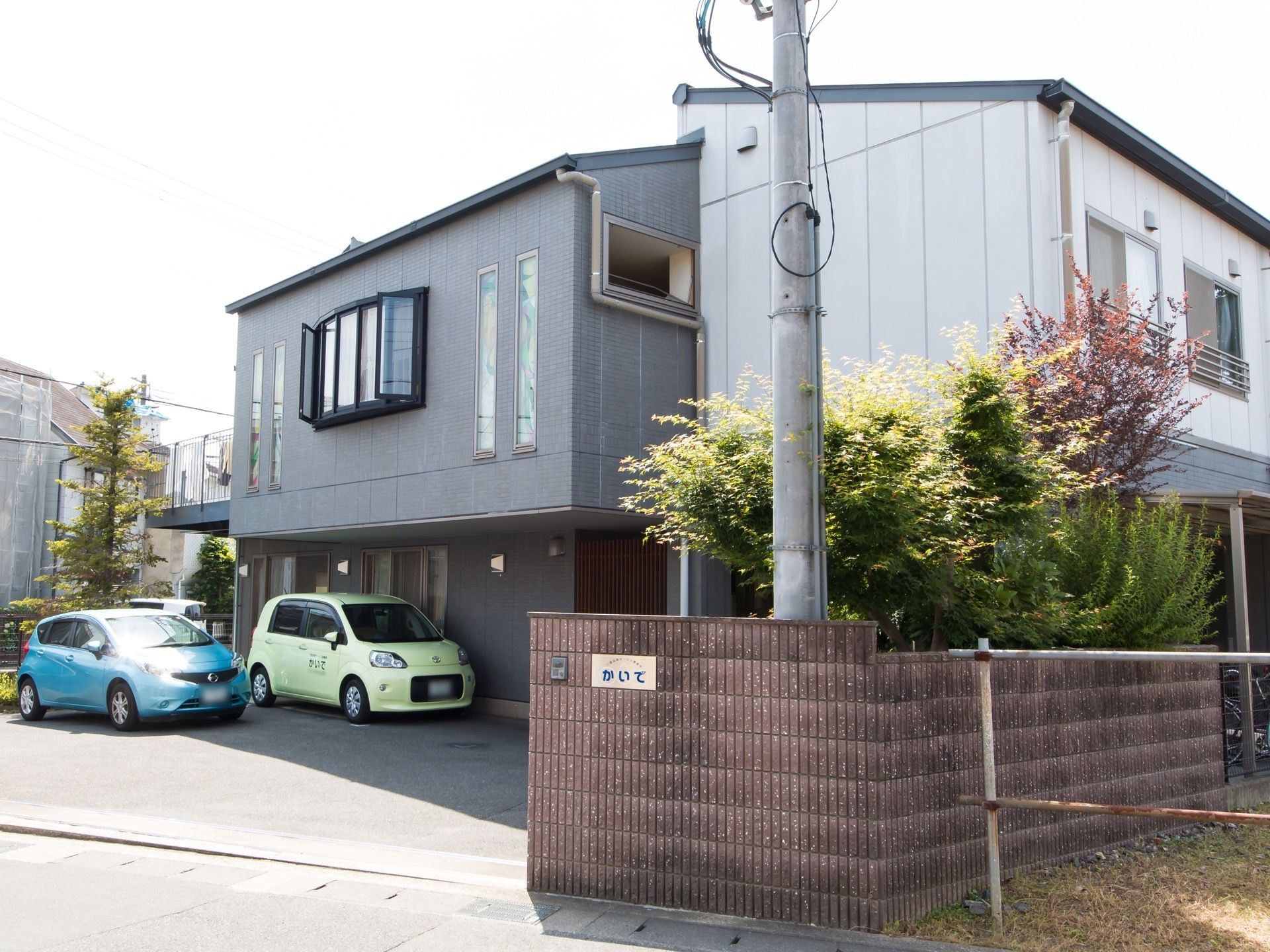 建物は、グレーと白の外壁の部分がある。駐車場の入口に塀があり、看板が設けられている。そしてインターフォンも付いている。